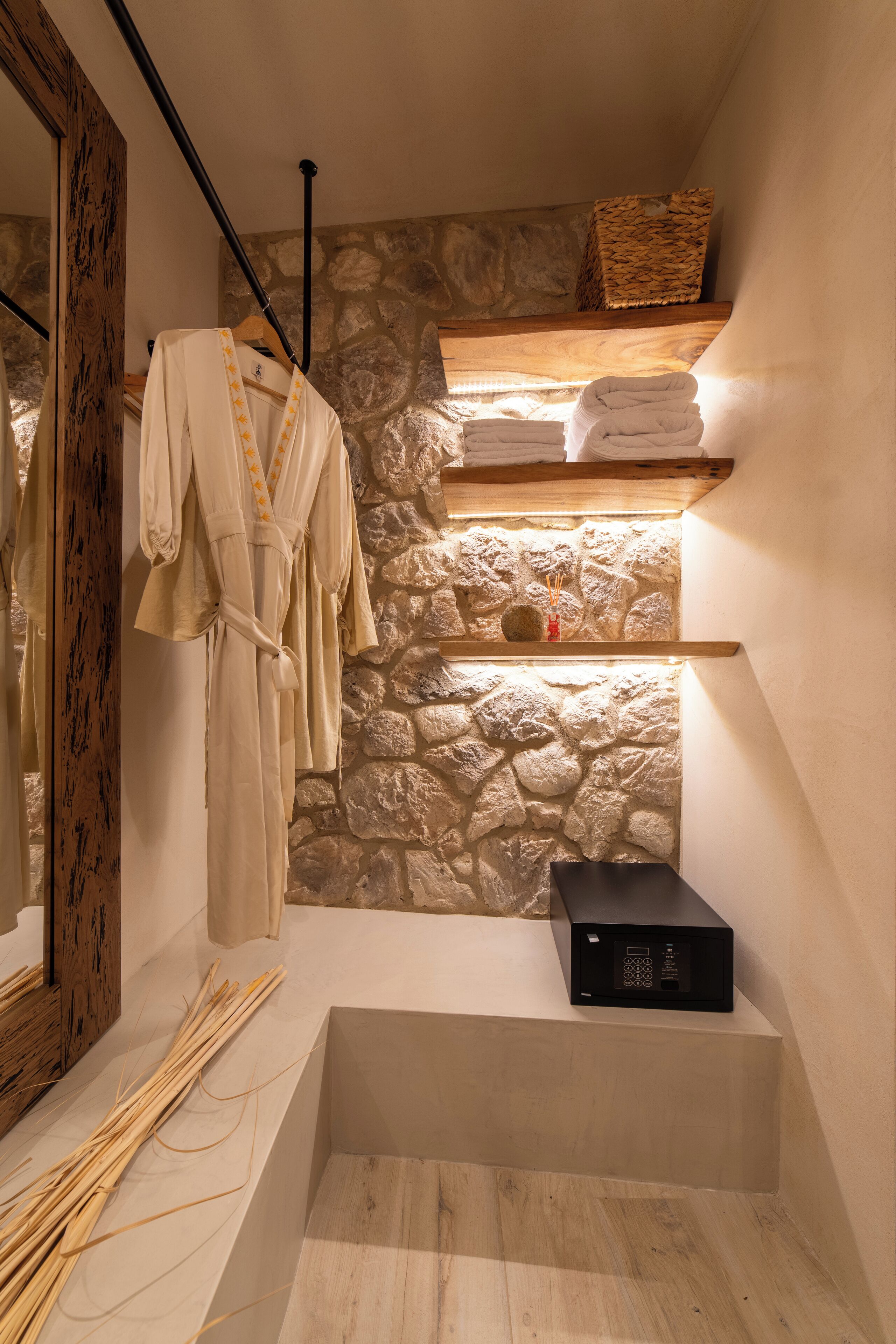 Une salle de bain avec un mur en pierre, une étagère en bois avec des serviettes, une robe suspendue, un miroir et un appareil noir posé sur une étagère.