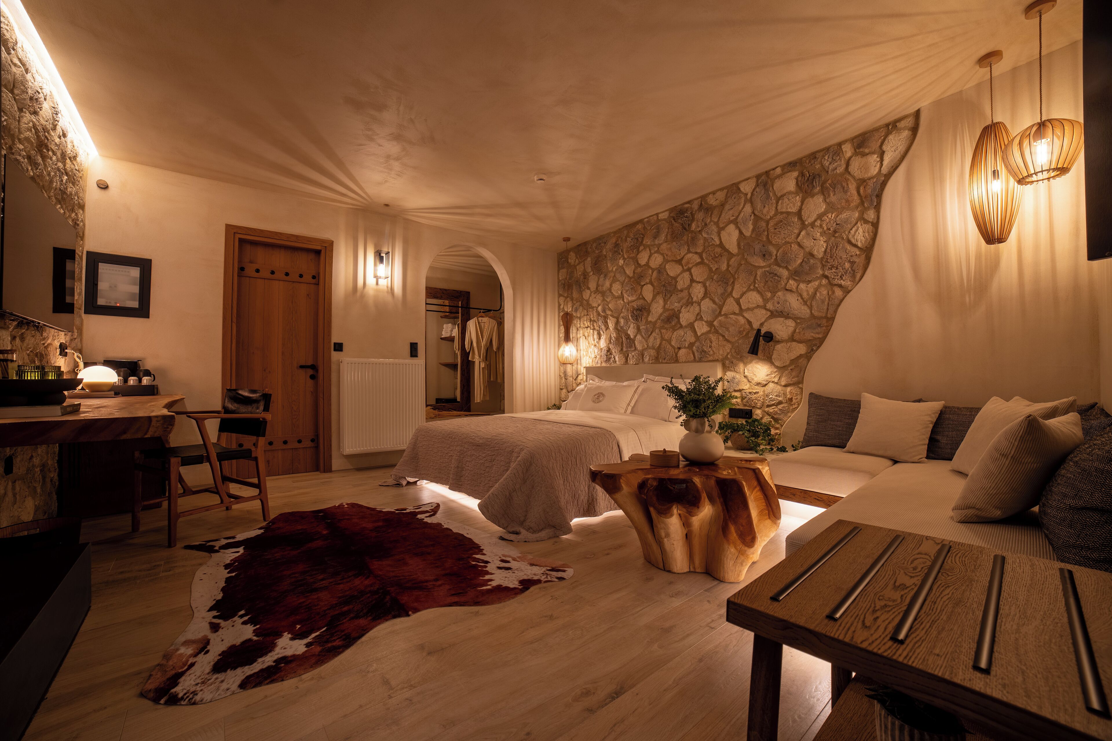 Een knus slaapkamer met een stenen muur, een houten bed, een bureau met stoel en een kleden van koeienhuid.