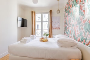 A hotel room with a bed, a flat-screen TV, a window with curtains, and a wall with a tropical print.
