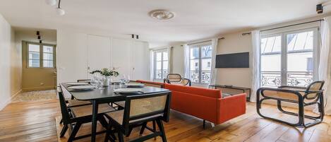 A modern living room with a dining table set for four, a sofa, a chair, and a TV on the wall.
