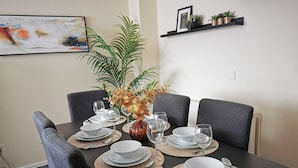 Una mesa de comedor dispuesta con platos, cuencos y cubiertos blancos, rodeada de sillas tapizadas en gris.