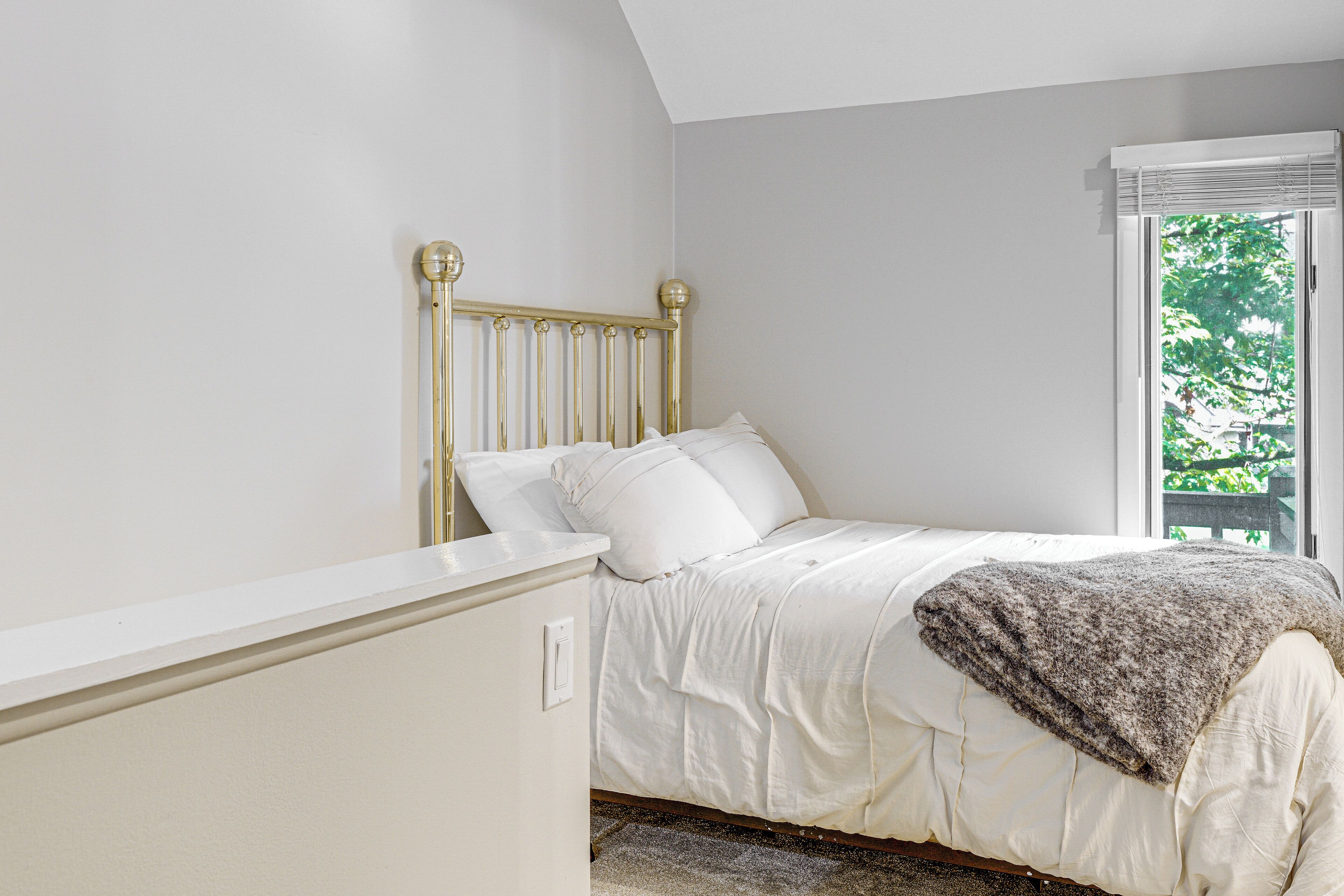 A bedroom with a bed, white walls, a window with blinds, and a view of greenery outside.