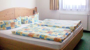 A double bed with a wooden headboard, two pillows, and a quilt. There is a window with curtains and a radiator below it. - Holiday apartment with balcony (Längenfeld)