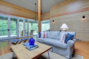A cozy living room with a wooden ceiling, a striped sofa, a coffee table with books, and large windows offering a view of greenery. - Serene Washington Island Getaway w/ Private Beach! (Washington Island)