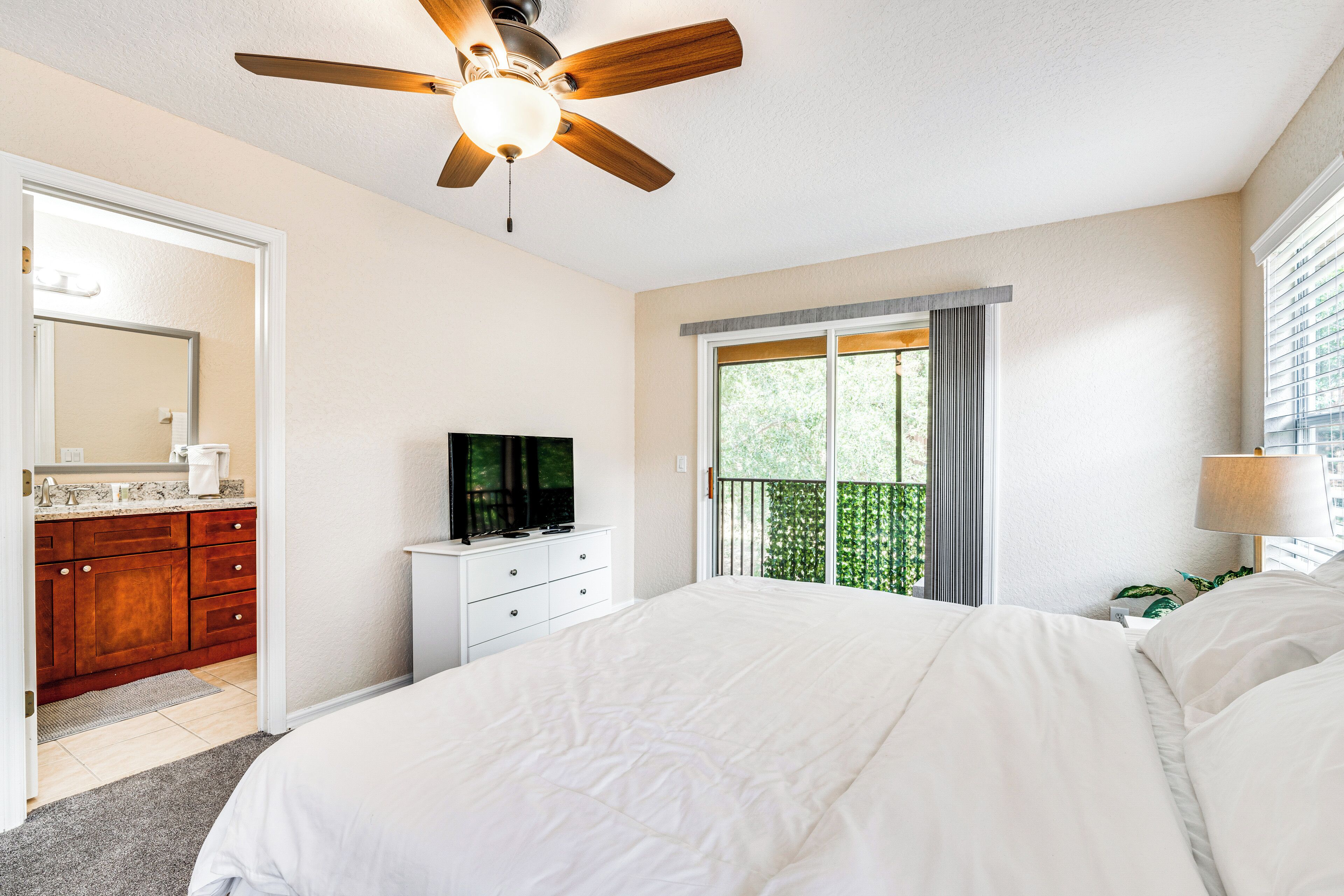 A bedroom with a bed, a ceiling fan, a TV, a window with curtains, and a bathroom with a vanity and mirror.
