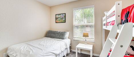 A bedroom with a bunk bed, a small bedside table, a window with blinds, and a single bed with a pillow.
