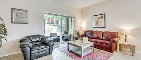 A living room with a black leather sofa, a red leather sofa, a wooden coffee table, and a floor lamp. There is a large window with a view of trees outside.