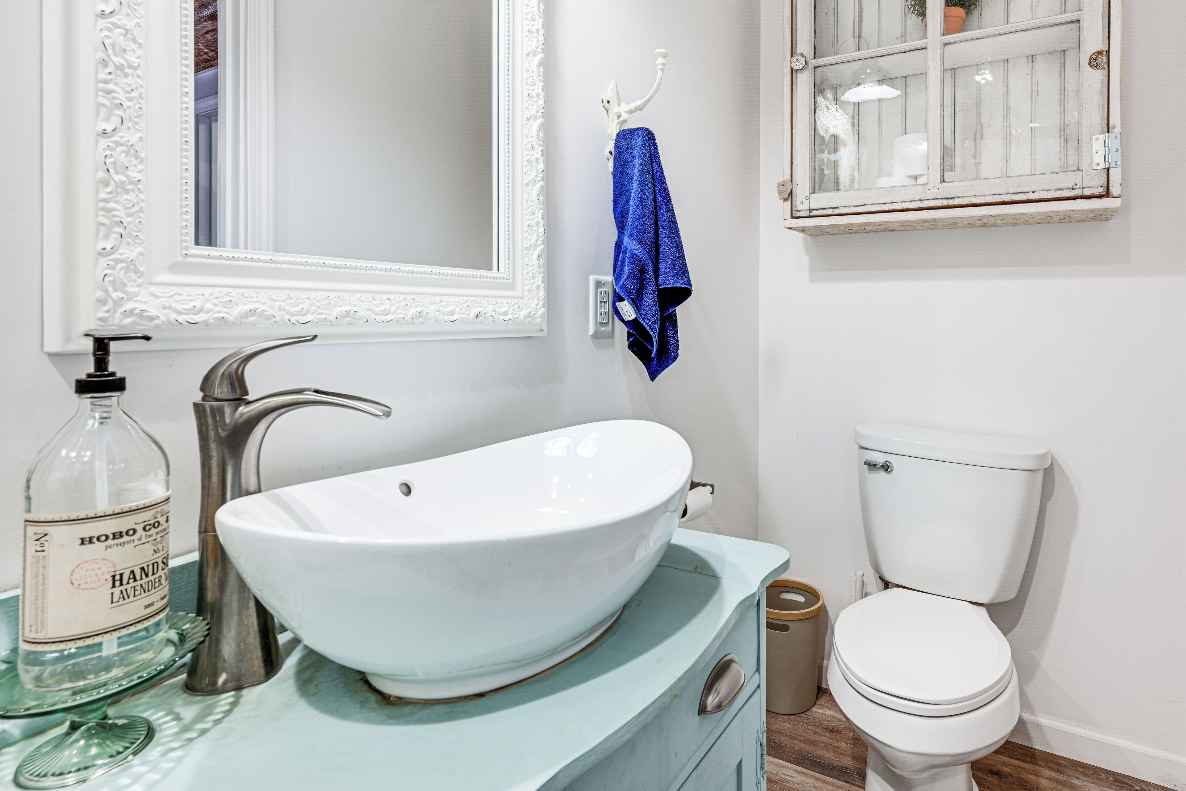 A bathroom with a white sink, a vintage-style faucet, a glass bottle, a blue towel, a toilet, and a mirror with an ornate frame.