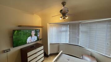 A bedroom with a bed, a TV mounted on the wall, a ceiling fan, and a window with blinds.
