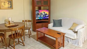 A living room with a wooden dining table, chairs, a white sofa, and a TV stand with a flat-screen TV. - Walkable Mobile Apartment w/ Fenced-in Yard (Mobile)