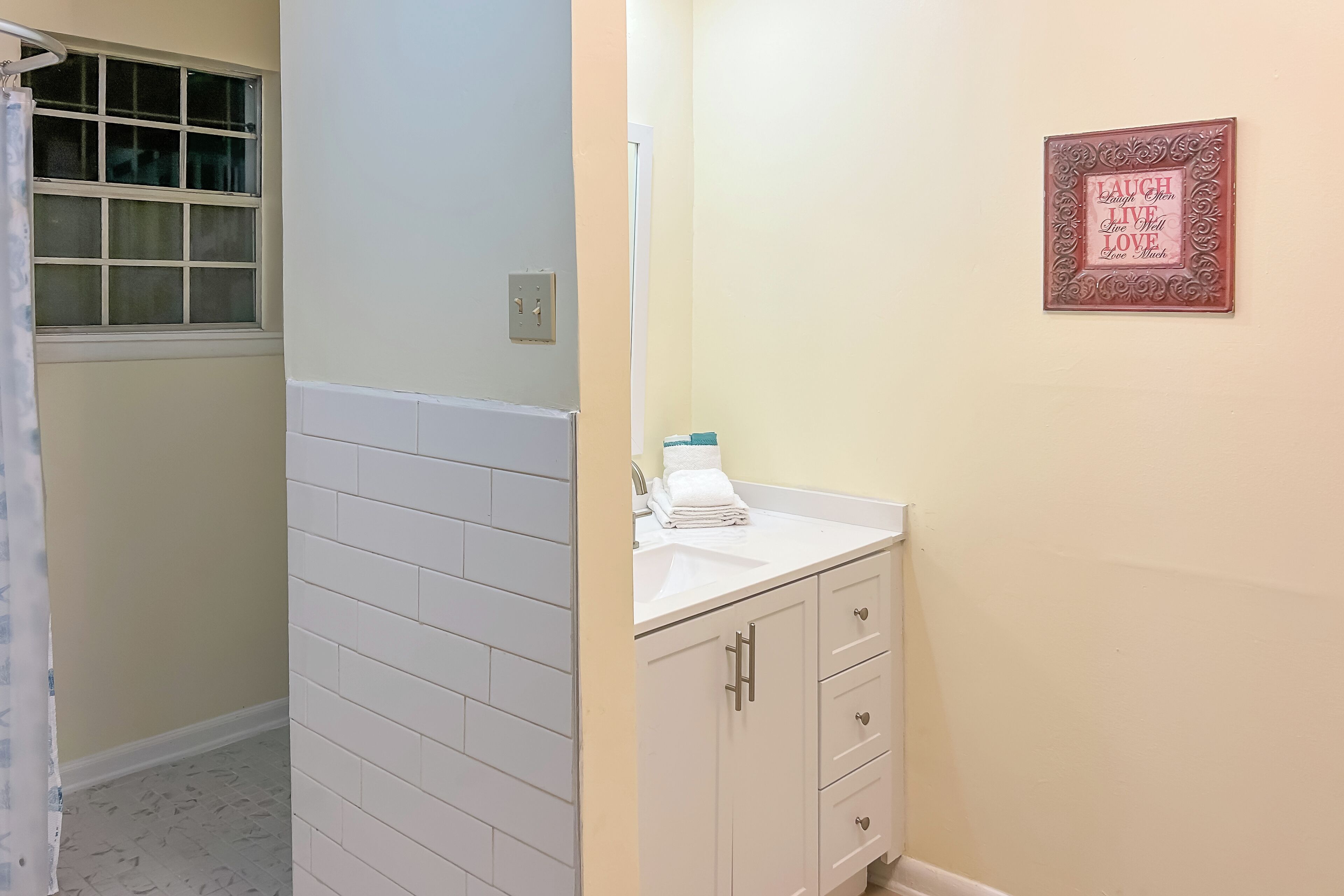 Een badkamer met een witte betegelde douche, een wastafel met een handdoek, en een gemotiveerd citaat in een lijst op de muur.