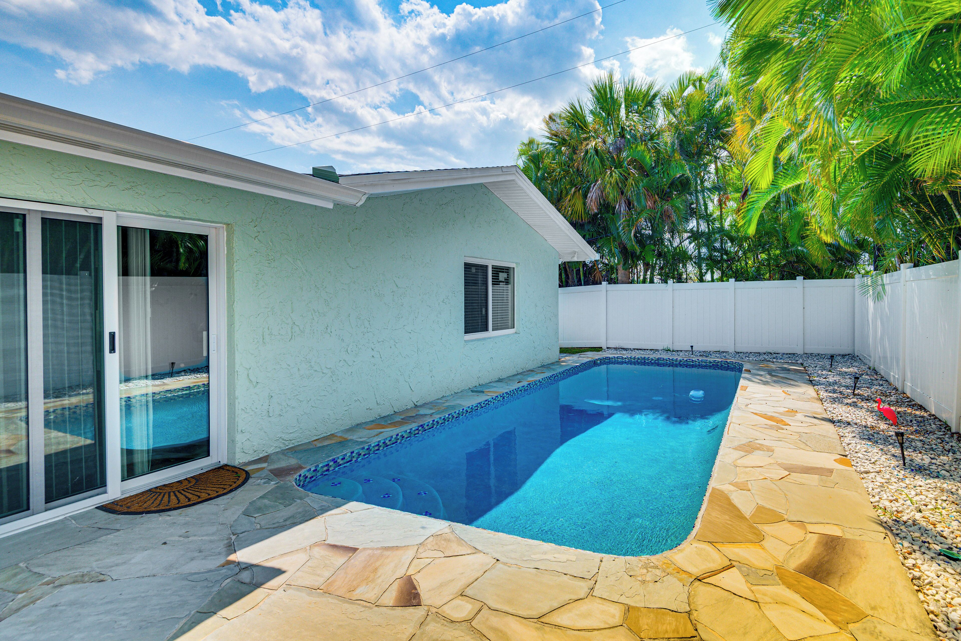 A swimming pool with stone tiles, a sliding glass door, and a white fence.