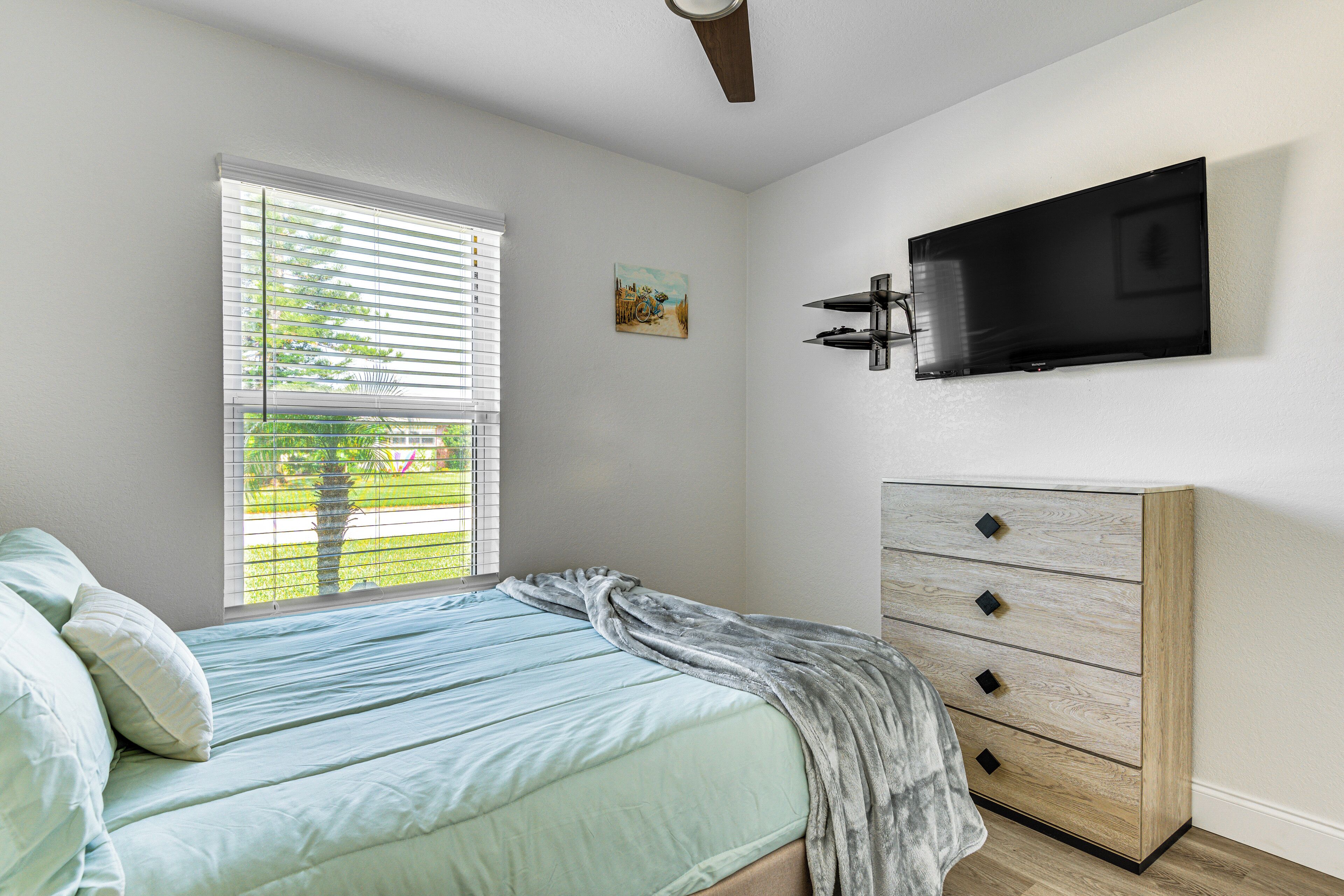 A bedroom with a bed, a dresser, a TV mounted on the wall, and a window with blinds.