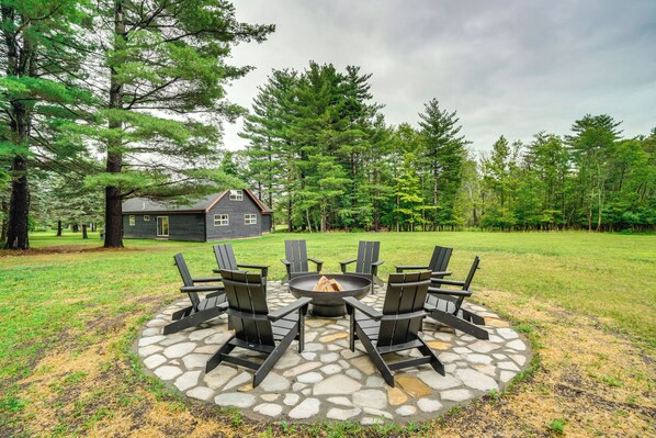 Een vuurplaats van natuursteen met zes Adirondack-stoelen eromheen geplaatst, gelegen in een grasveld met een huis op de achtergrond.