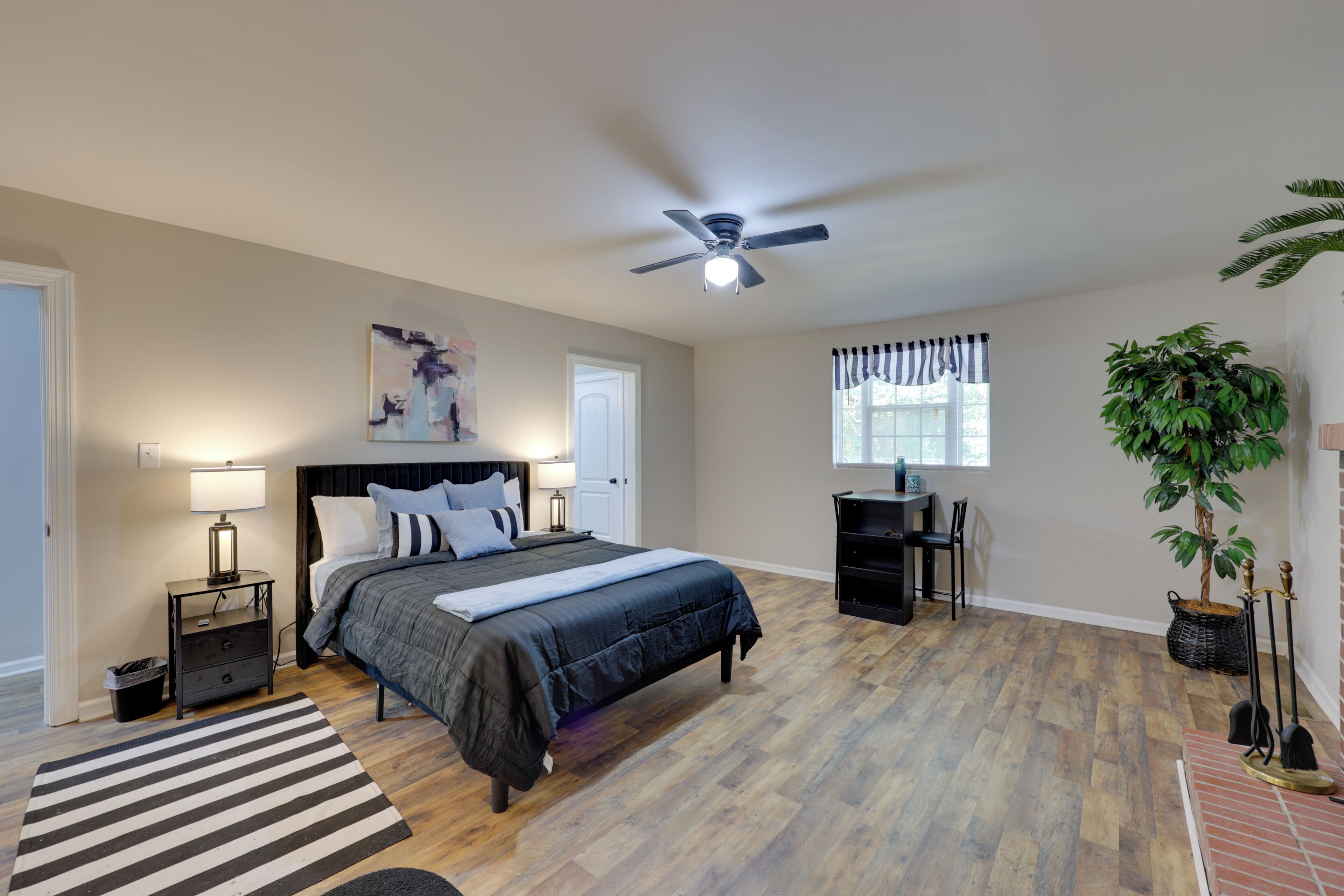 A bedroom with a bed, nightstands, a ceiling fan, a window with curtains, a plant, and a fireplace.