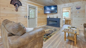 Interior acogedor de una cabaña con techo de madera, un televisor sobre la chimenea, un sofá con un cojín estampado y un tablero de ajedrez en una mesa de madera.