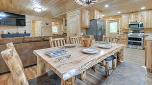 Interior rústico de una cabaña con una mesa de comedor de madera, sillas, un sofá, una cocina con electrodomésticos y un televisor montado en la pared.