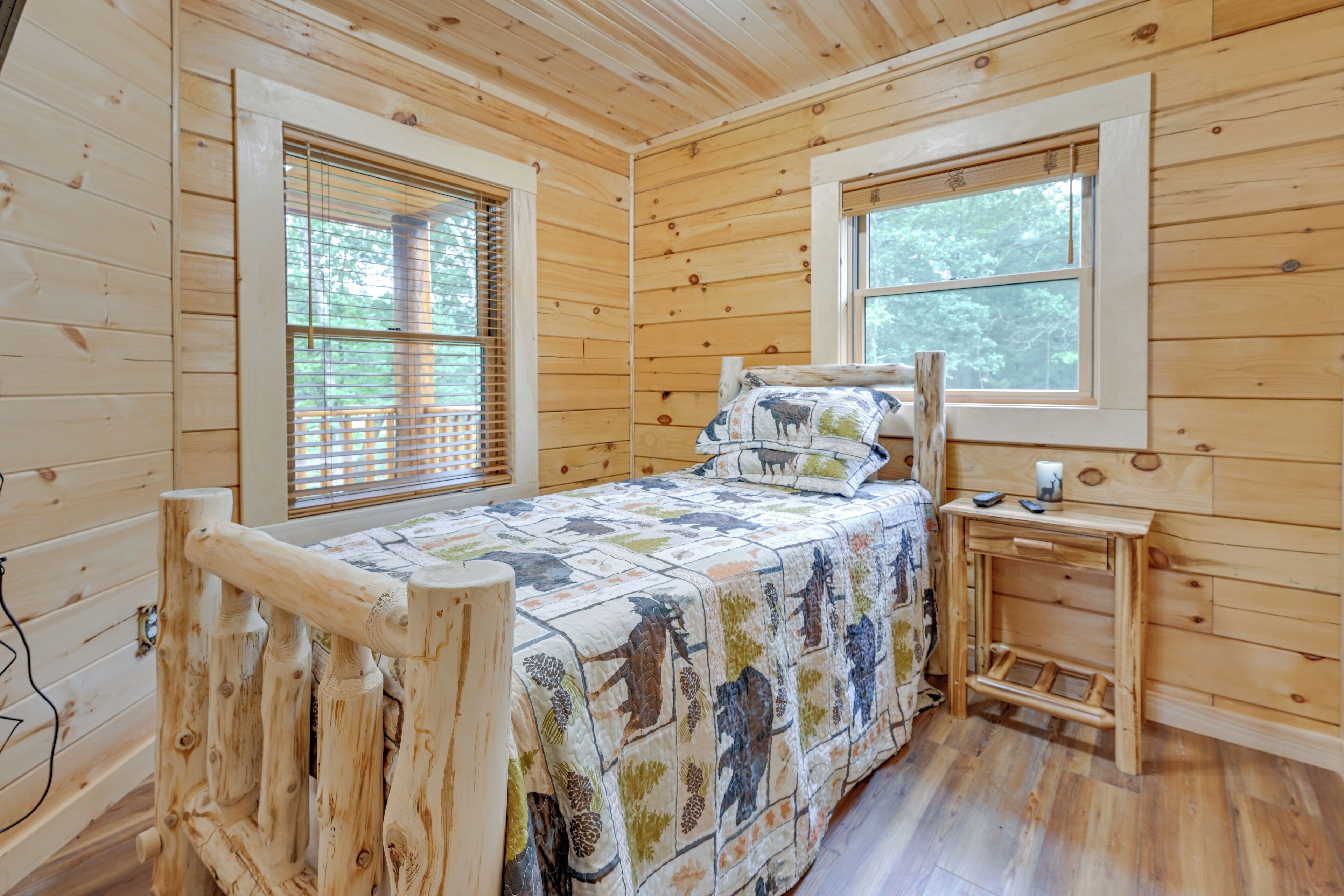 A wooden cabin bedroom with a bed, two windows, and a small wooden nightstand.