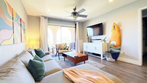 Un salon moderne avec un canapé, une table basse et un téléviseur. On y trouve également un ventilateur de plafond, une planche de surf et un balcon avec vue.