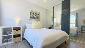 Une chambre moderne avec un grand lit, une table de chevet, un petit bureau et une bibliothèque. Un grand miroir est accroché à un des murs, et un tableau encadré se trouve au-dessus du lit.