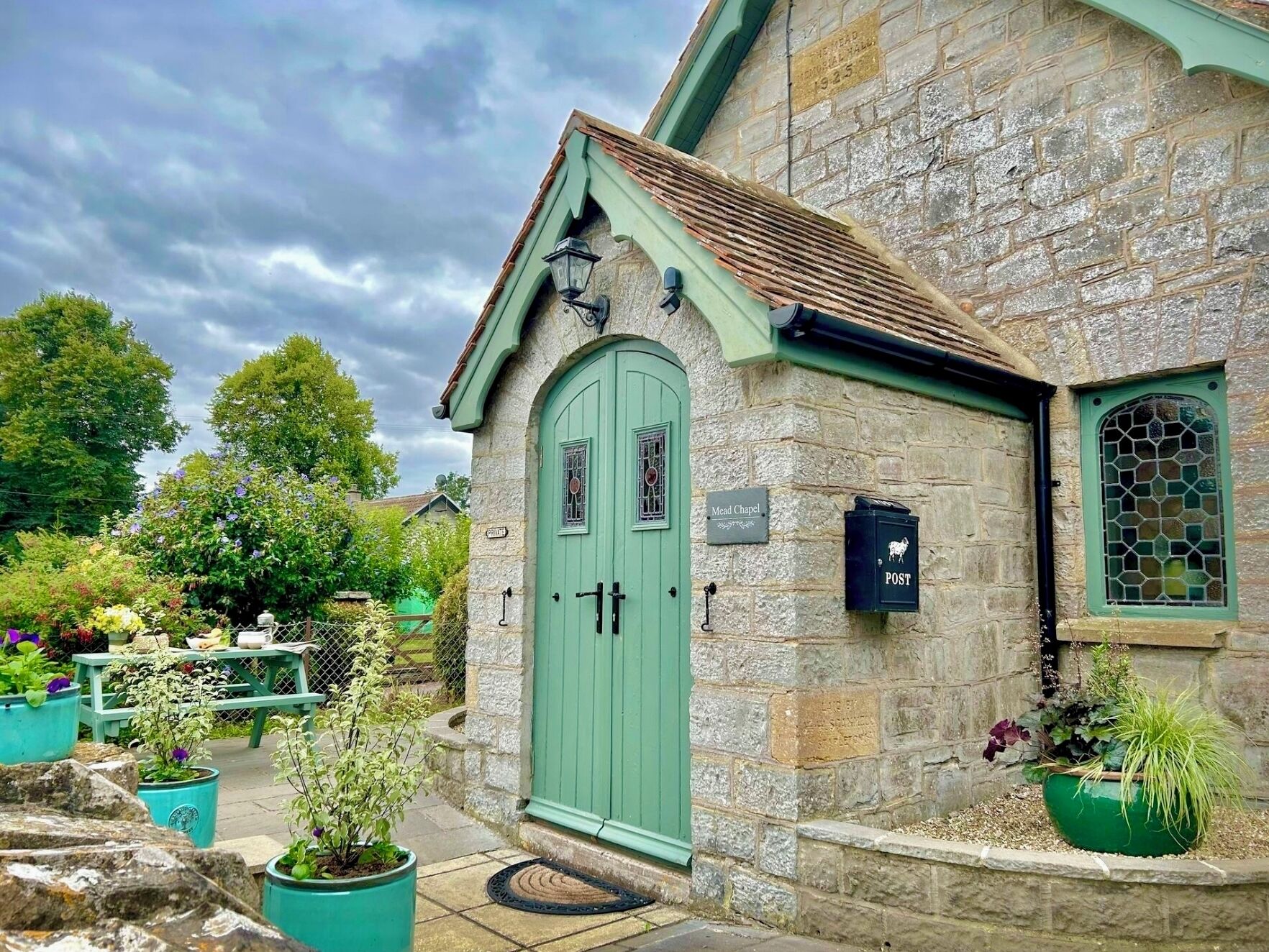 A stone building with a green door, a sign for 'Mead Chapel', and a 'POST' sign.