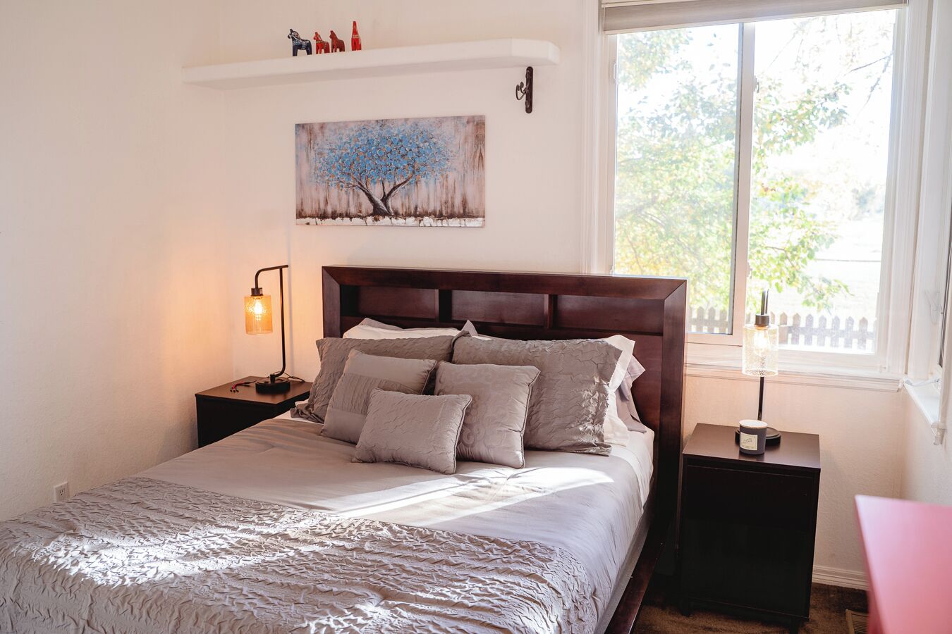 A neatly made bed with a textured blanket, a wooden headboard, and a painting above it. There are two bedside tables with lamps, and a window with a view of trees.