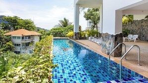 A swimming pool with blue mosaic tiles, surrounded by greenery and a stone wall. - Kata Ocean View 2BR Apartment O910 (Karon)