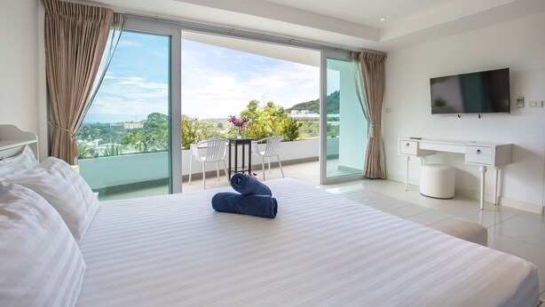 A modern hotel room with a large bed, a desk, a flat-screen TV, and a balcony with a view. - Kata Ocean View 2BR Apartment O910 (Karon)