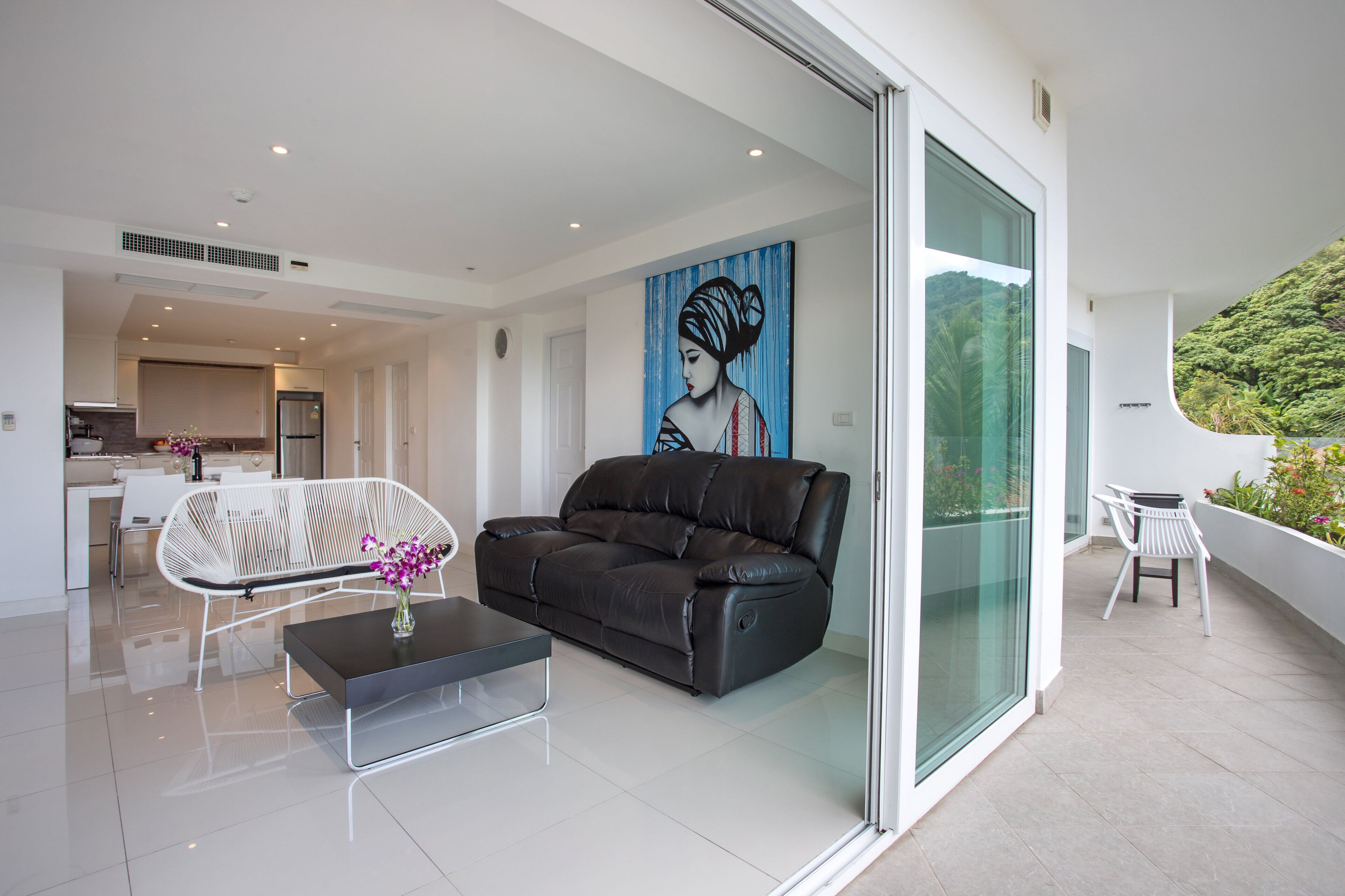 Un salon moderne avec un canapé en cuir noir, un fauteuil blanc et une table basse. Un tableau orne le mur, et une porte vitrée donne accès à un balcon d’où l’on peut admirer la verdure.