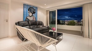 A modern living room with a black leather sofa, a white chair, and a glass coffee table. There is a painting on the wall and a view of the city through the balcony doors. - Kata Ocean View 2BR Apartment O910 (Karon)