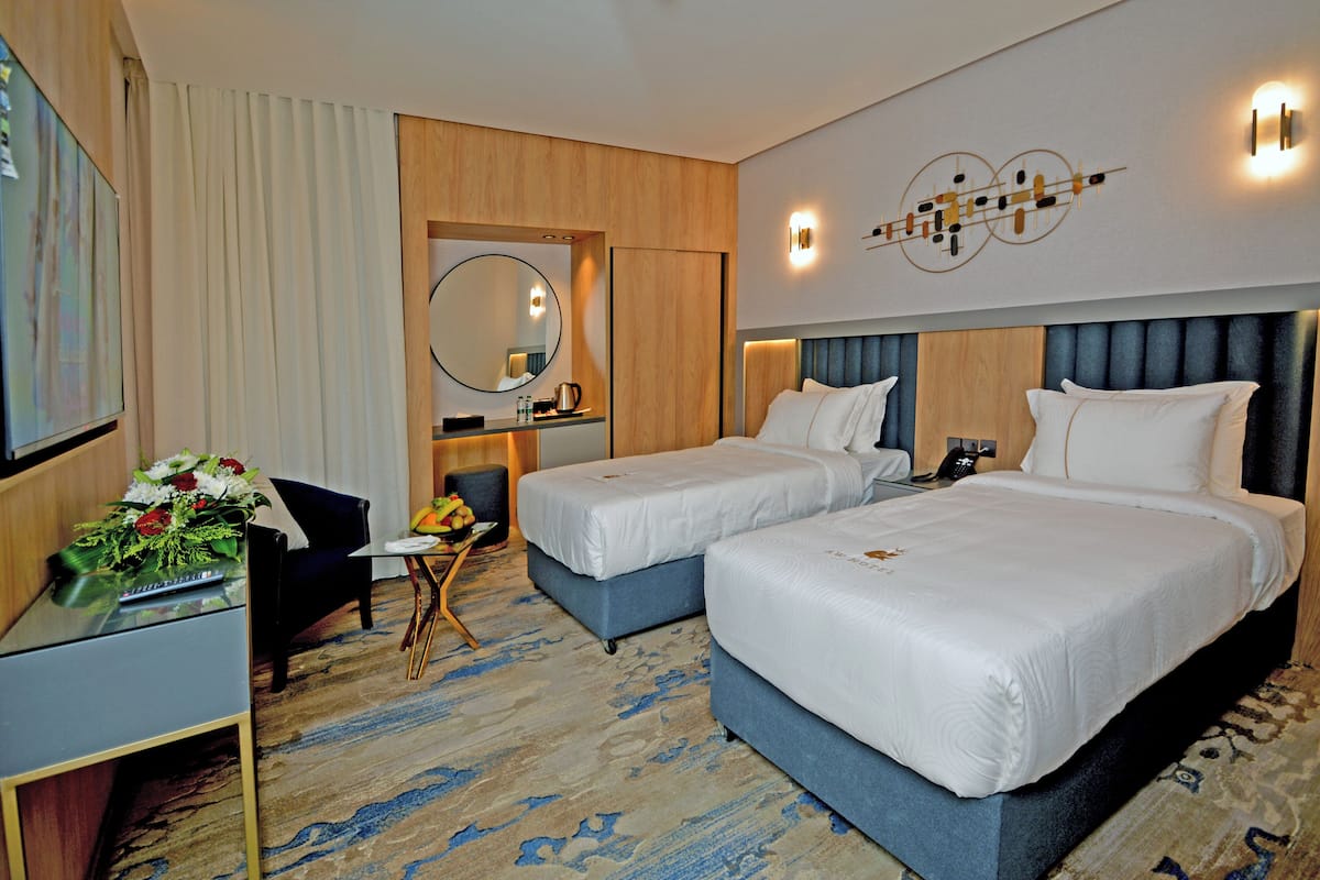 A hotel room with two beds, a desk, a chair, a small table with a vase of flowers, and a mirror.