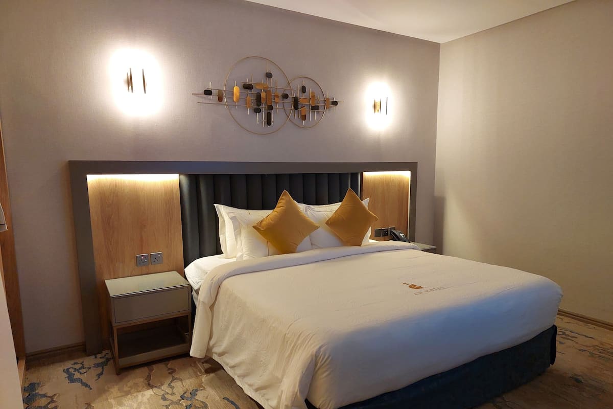 A hotel room with a large bed, a nightstand, a wall-mounted light fixture, and a carpeted floor.