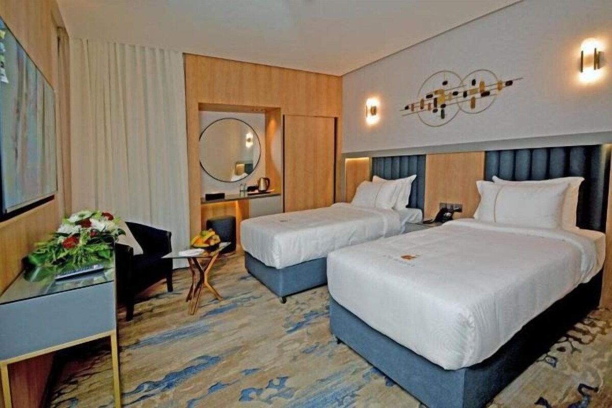 A hotel room with two beds, a desk, a chair, and a small table with a vase of flowers.