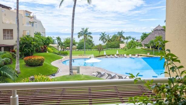 A resort pool area with a clear view of the ocean, surrounded by palm trees and a well-maintained lawn.