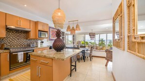 A modern kitchen with wooden cabinets, a granite countertop, and a central island. There are hanging pendant lights and a large window offering a view of the outdoors.