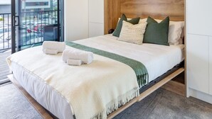 A modern hotel room with a large bed, a wooden headboard, and a view of the parking area through a glass door. - Upper Studio Perfection: 8 Wilmer St. Gem (Christchurch)