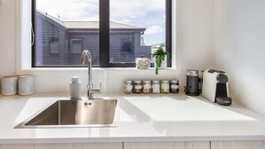 A kitchen with a stainless steel sink, a window with a view of a house, jars of condiments, and a coffee maker. - Upper Studio Perfection: 8 Wilmer St. Gem (Christchurch)