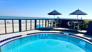 A swimming pool with a view of the beach, a deck area with chairs and umbrellas, and a lifeguard stand. - Beach Happy - Ocean View at Symphony Beach Club (Ormond Beach)