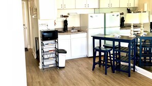 A compact kitchen with white cabinets, a microwave, and a coffee maker. There is a dining area with a table and chairs. - Waters Edge - Ocean View at Symphony Beach Club (Ormond Beach)