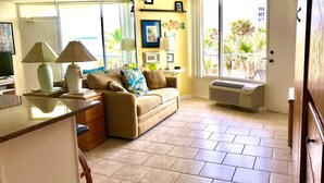 A living room with a beige sofa, a coffee table, and a TV. There is a window with a view of palm trees and a door leading outside.