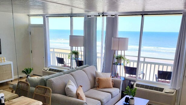 A modern living room with a large window overlooking the beach, a grey sofa, wooden flooring, and a small table with chairs. - The Lookout - Oceanfront at Symphony Beach Club (Ormond Beach)