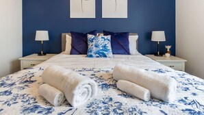 Un lit bien fait, recouvert d’une couette à motifs floraux, avec deux oreillers et deux serviettes enroulées, le tout placé devant un mur bleu orné de deux œuvres d’art encadrées.