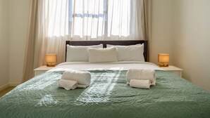 A neatly made bed with a green quilt, two bedside lamps, and a window with sheer curtains.