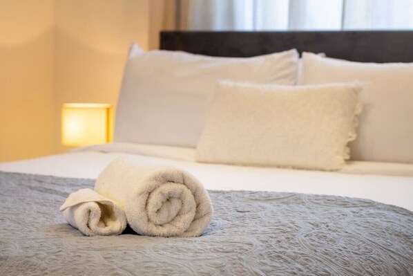 A neatly made bed with a rolled white towel, a textured gray bedspread, and a bedside lamp.