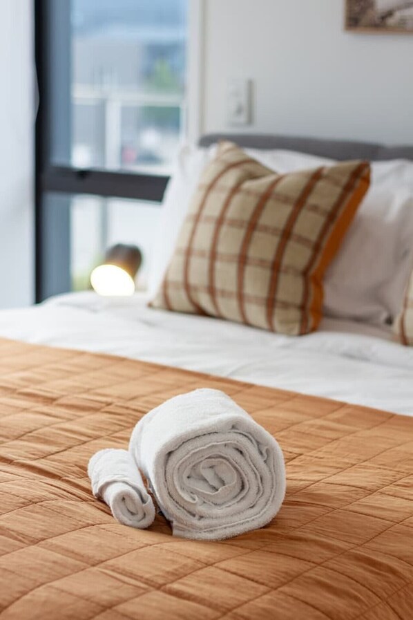 Una cama bien tendida con una toalla blanca enrollada y un cojín estampado en pata de gallo.