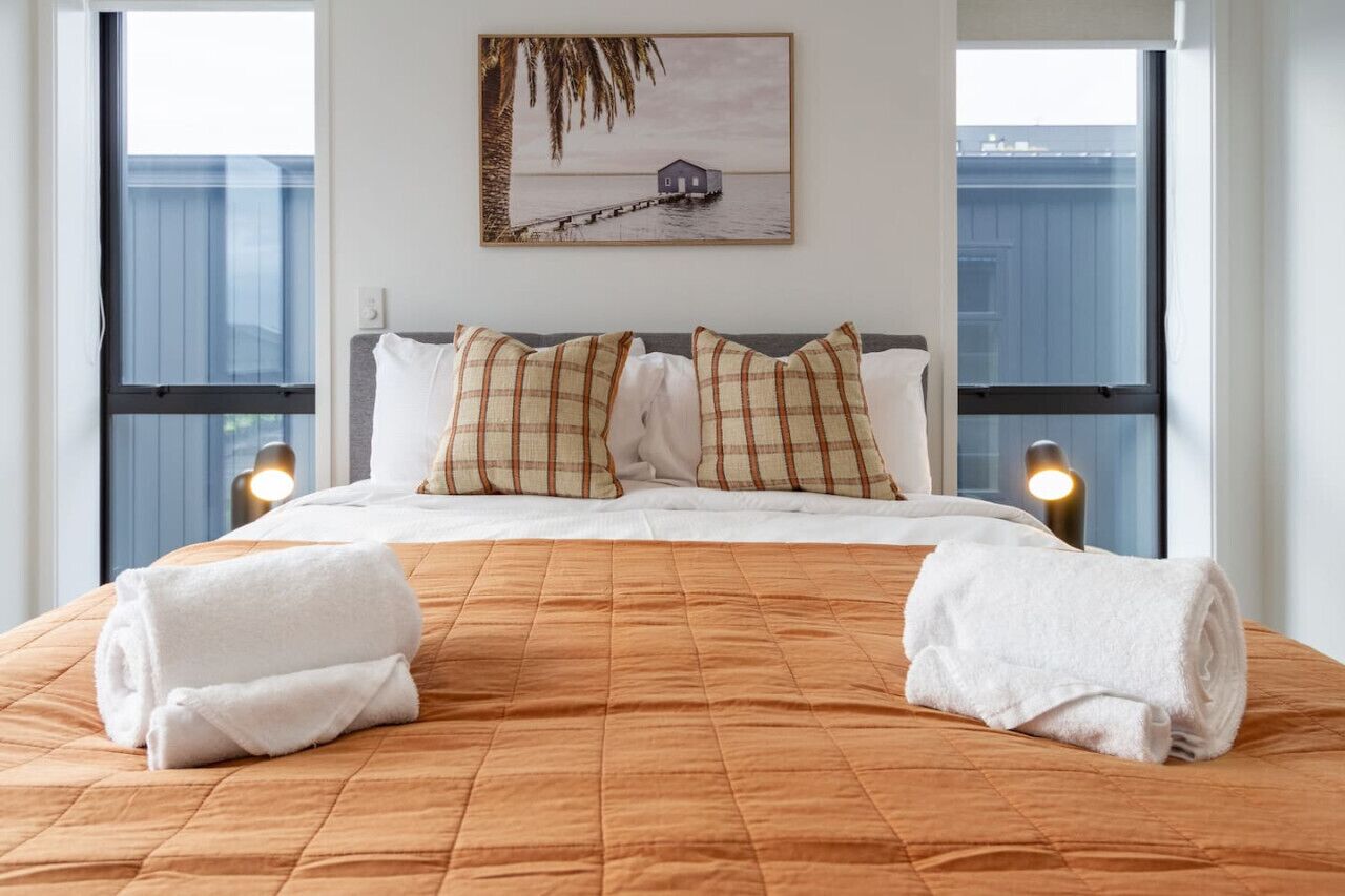 A neatly made bed with a brown quilt, two white towels, and plaid pillows. A framed picture of a house on stilts over water hangs on the wall.