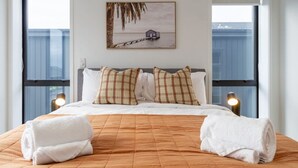 Una cama bien hecha con un edredón marrón, dos toallas blancas y almohadas a cuadros. Un cuadro enmarcado de una casa sobre pilotes en el agua cuelga en la pared.