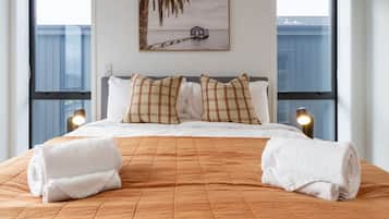 A neatly made bed with a brown quilt, two white towels, and plaid pillows. A framed picture of a house on stilts over water hangs on the wall.
