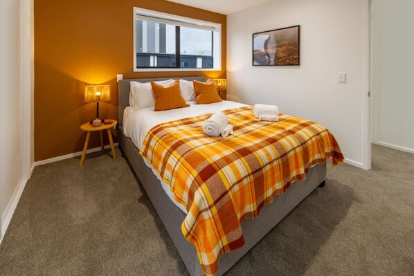A bedroom with a bed, a window, a wall-mounted picture, and a nightstand with a lamp.