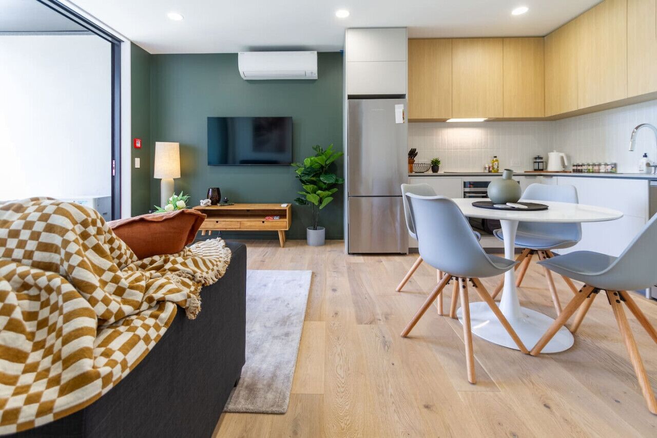 A modern hotel room with a sofa, a dining table with chairs, a kitchen area with a refrigerator, and a flat-screen TV mounted on the wall.